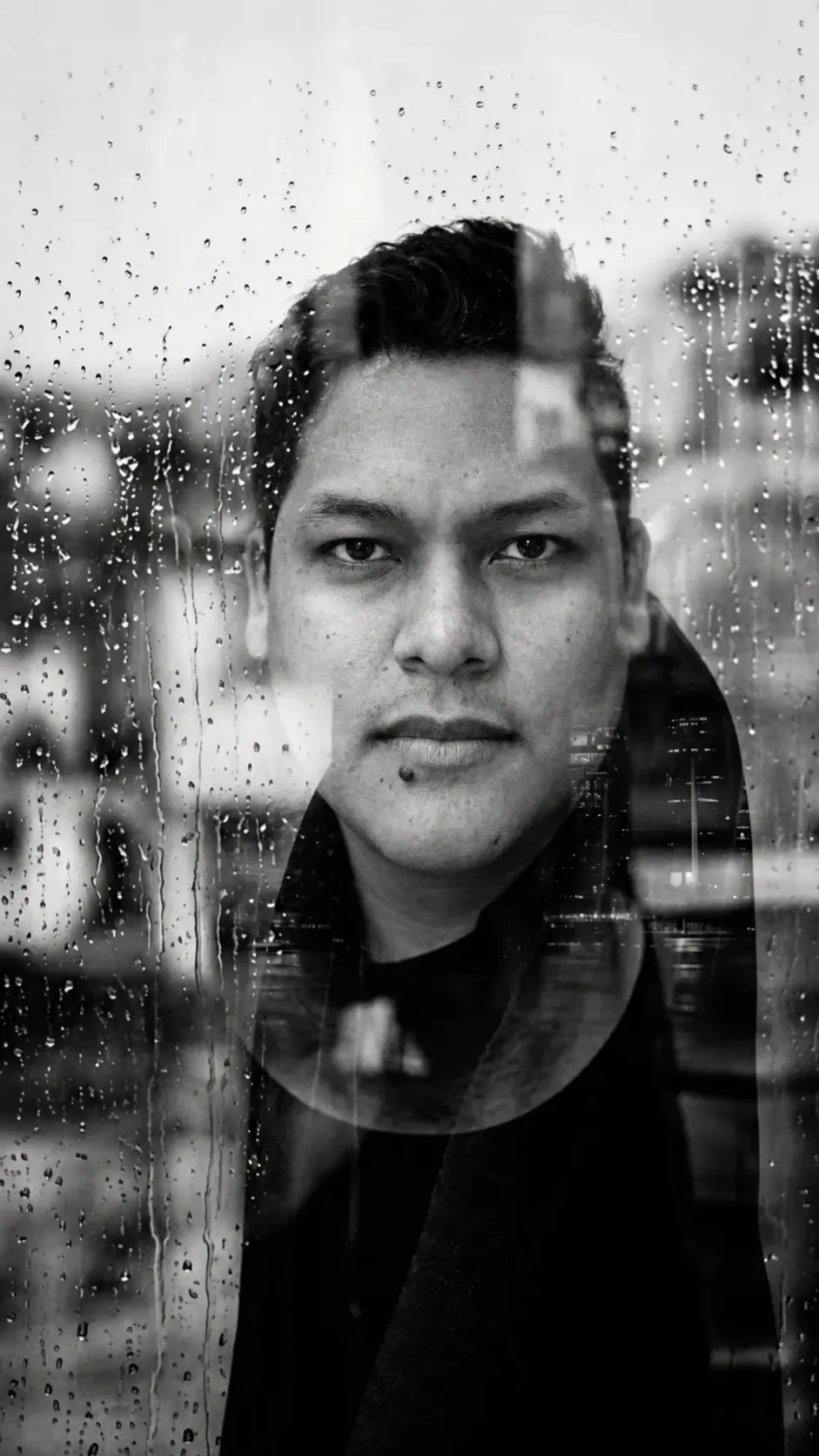 Moisés Mota detrás de una ventana con lluvia, con la ciudad reflejada
